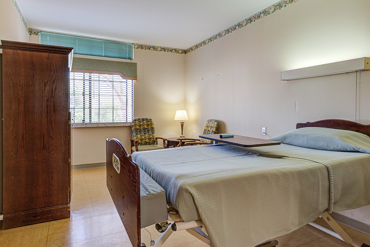 Single occupancy room with bed, chairs, cabinet and window.