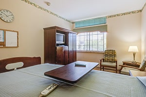 Resident room with blue bedding, large window, TV and chairs and a clock on the wall